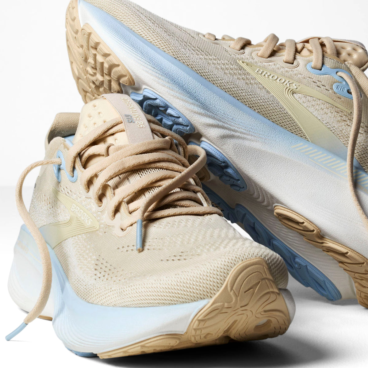 A pair of beige and light blue running shoes with textured soles, mesh fabric, and laces, positioned on a white background, with one shoe resting on top of the other.