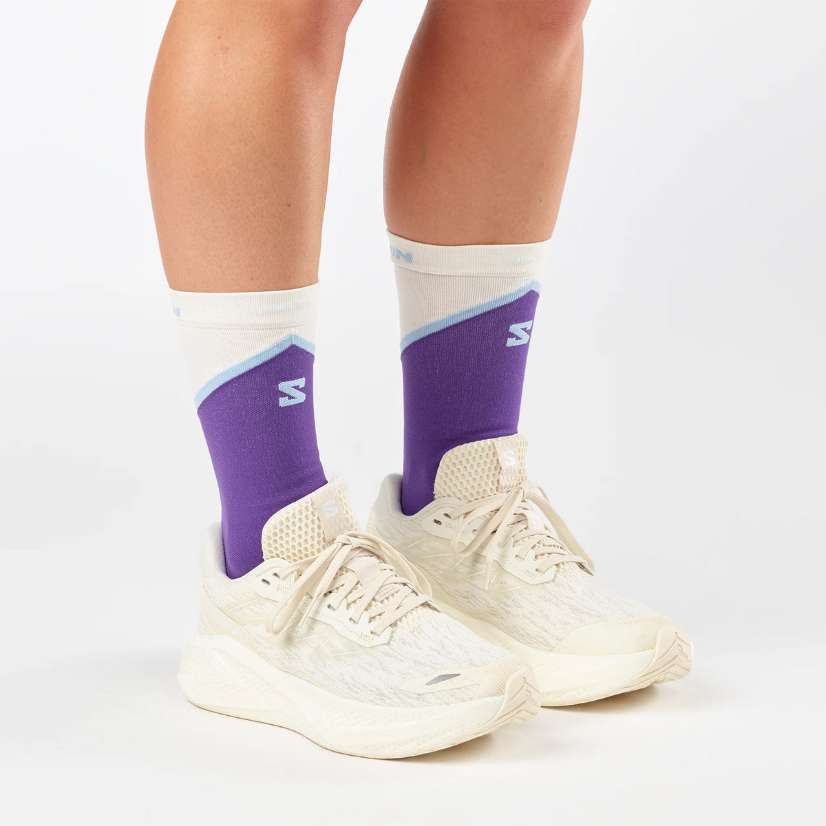Person wearing cream-colored athletic shoes and cream, blue, and purple crew socks with a white S logo, standing on a white background. Only the lower legs and feet are visible.