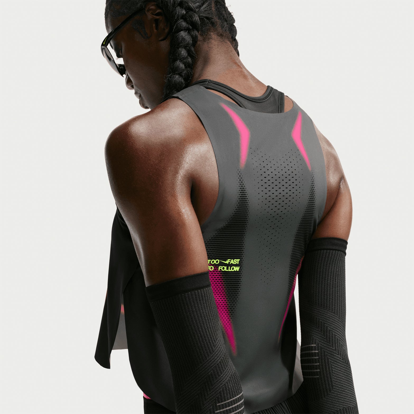 A runner with braided hair and sunglasses faces away, showcasing the advanced details of the Nike Women’s Dri-FIT ADV Cropped Running Vest in Iron Grey / Volt / White.
