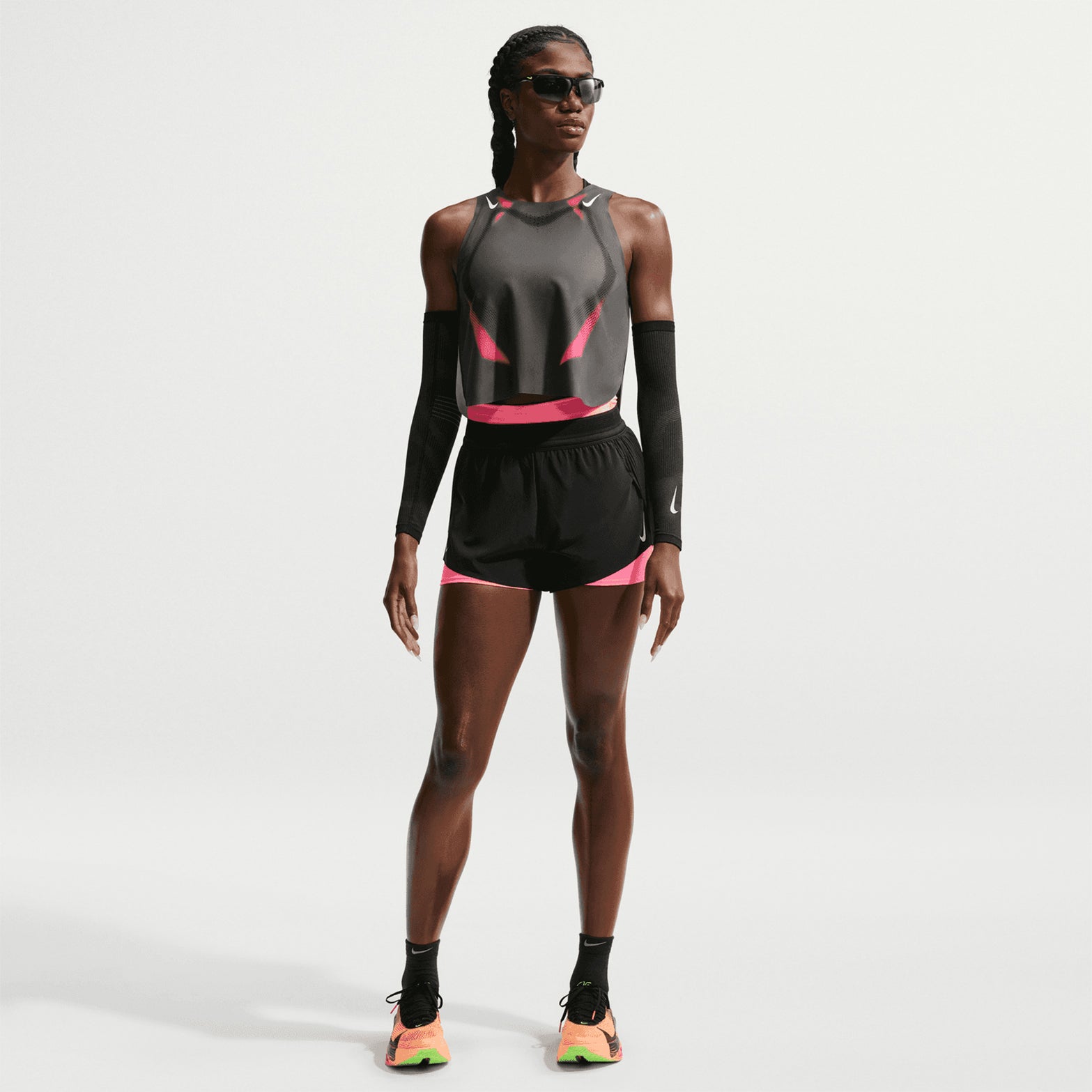 A person wearing a Nike Women’s Dri-FIT ADV Cropped Running Vest in Iron Grey, Volt, and White, paired with sunglasses and sneakers, stands against a plain white background.