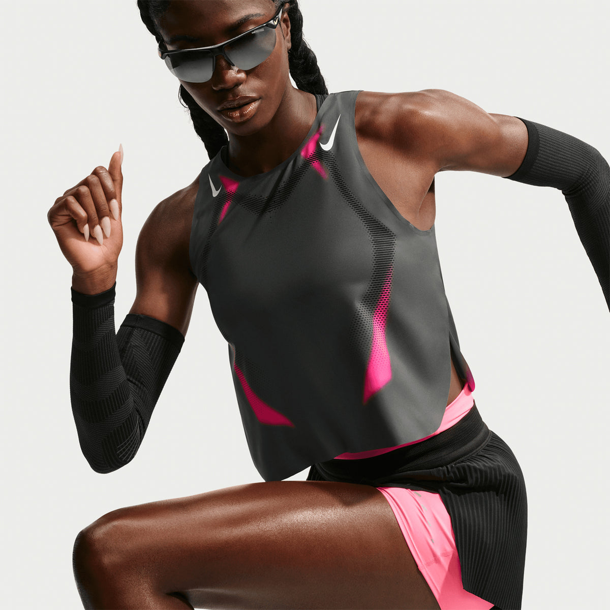 Athletic woman in black and pink sportswear and sunglasses poses in a running stance against a plain white background. She wears a sleeveless top, arm sleeves, and shorts.