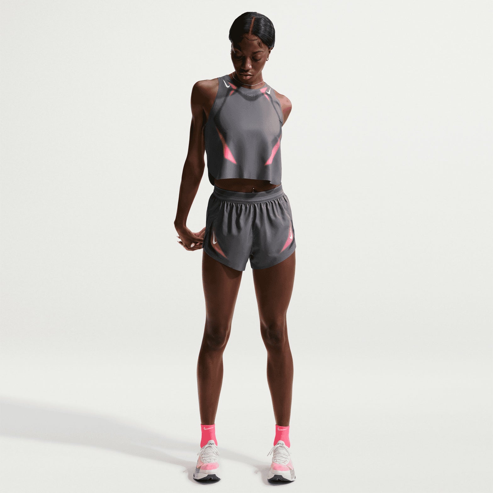 A person wearing a gray athletic outfit with pink accents, pink socks, and white sneakers stands on a white background, looking down with their hands by their sides. Their look is completed with the Nike Women’s Dri-FIT Aeroswift ADV 3 Inch Running Shorts in Iron Grey/White—lightweight performance shorts designed for comfort and style.