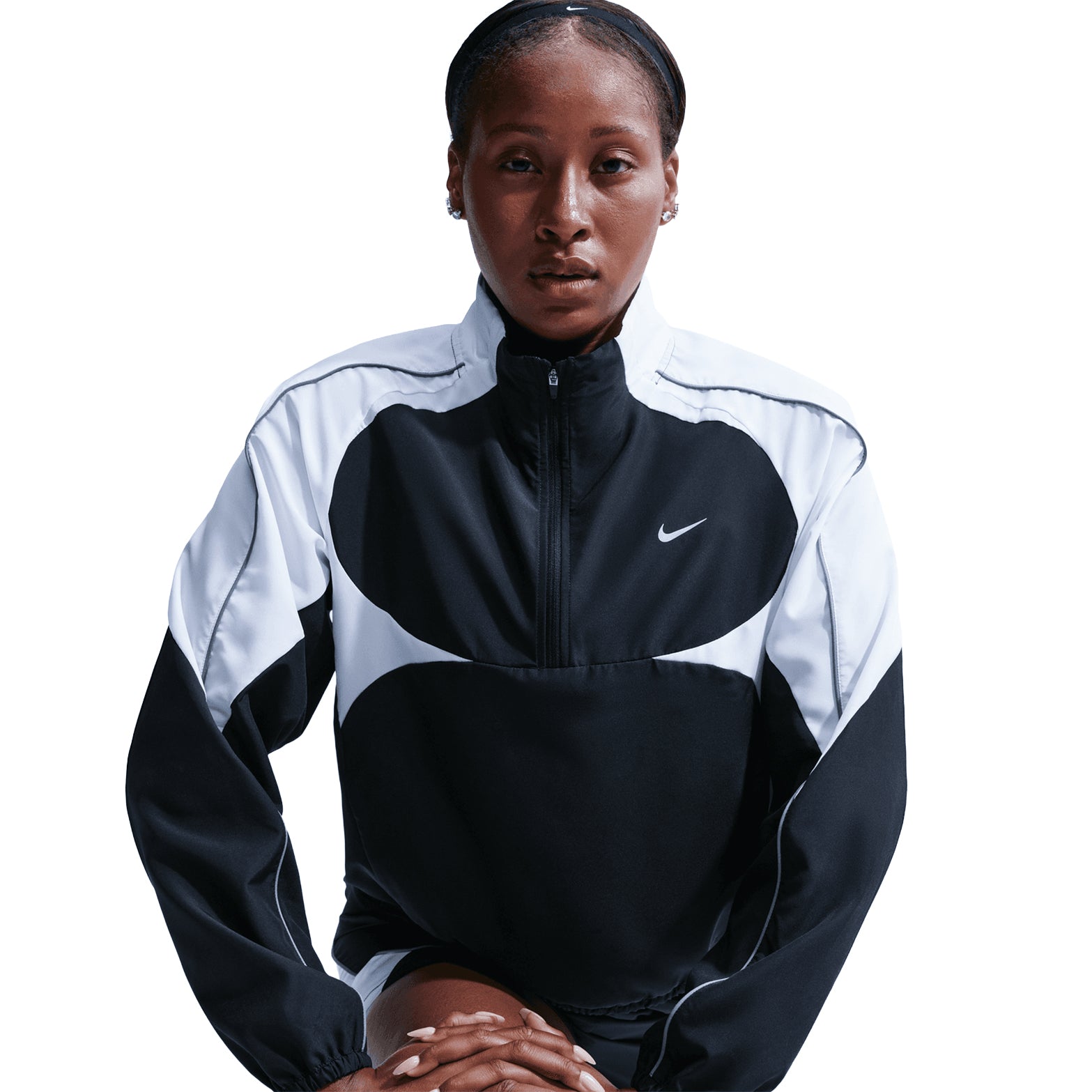 A woman wearing the Nike Women’s Dri-FIT Tempo Swoosh 1/2 Zip Running Jacket in Black, White, and Cool Grey sits with her hands resting in front of her, looking directly at the camera against a plain white background.