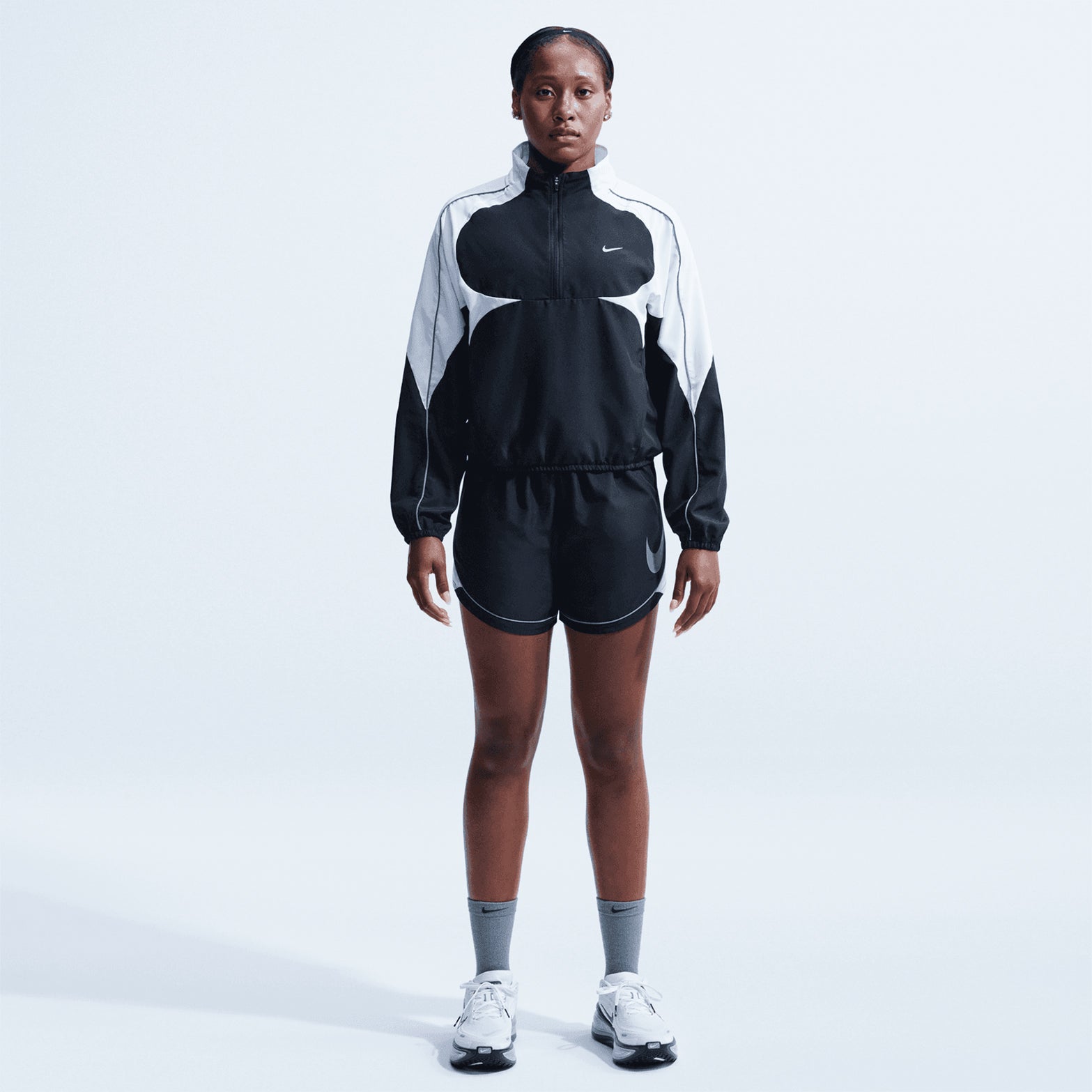 A person stands facing forward, wearing the Nike Women’s Dri-FIT Tempo Swoosh 1/2 Zip Running Jacket in Black, White, and Cool Grey, paired with matching shorts, gray socks, and white sneakers against a plain light background.