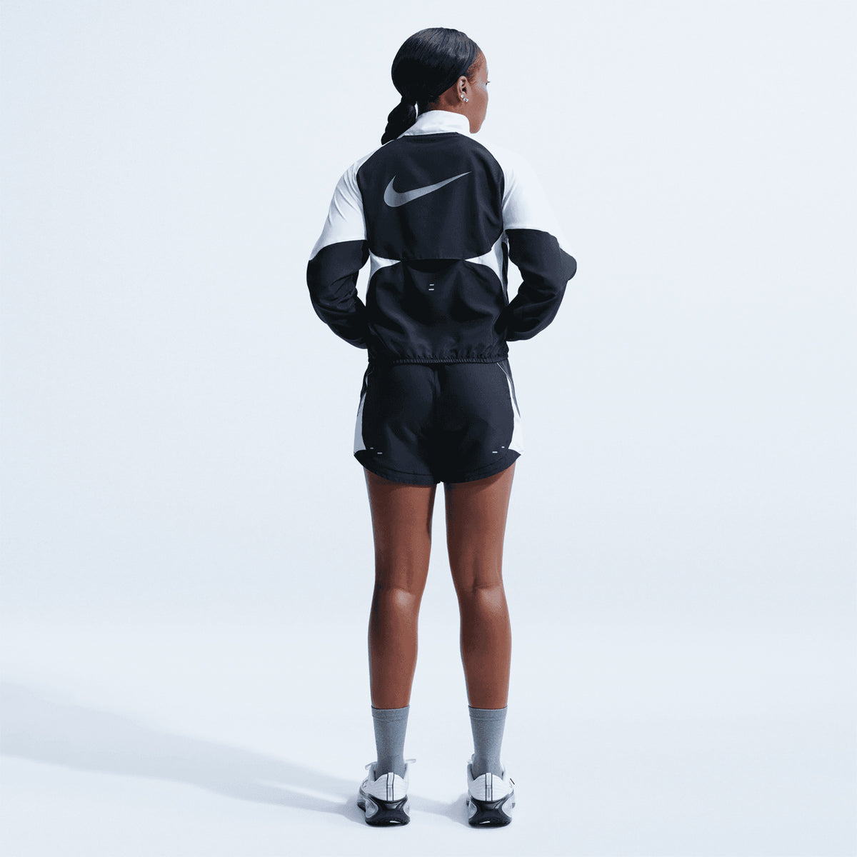 A person stands facing away, wearing the Nike Women’s Dri-FIT Tempo Swoosh 1/2 Zip Running Jacket in Black, White, and Cool Grey, with matching shorts, gray socks, and white sneakers against a plain light background.