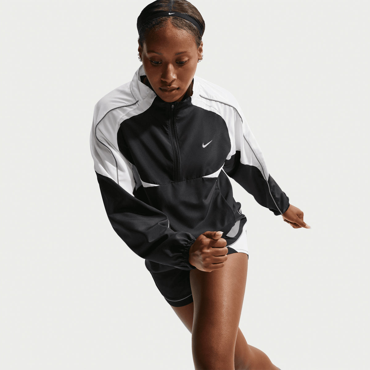 Woman in a black and white Nike athletic jacket and shorts appears to be running or in stride, set against a plain white background.