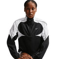 A person wearing the Nike Women’s Dri-FIT Tempo Swoosh 1/2 Zip Running Jacket in Black, White, and Cool Grey sits with hands resting on their knees, facing forward against a white background.