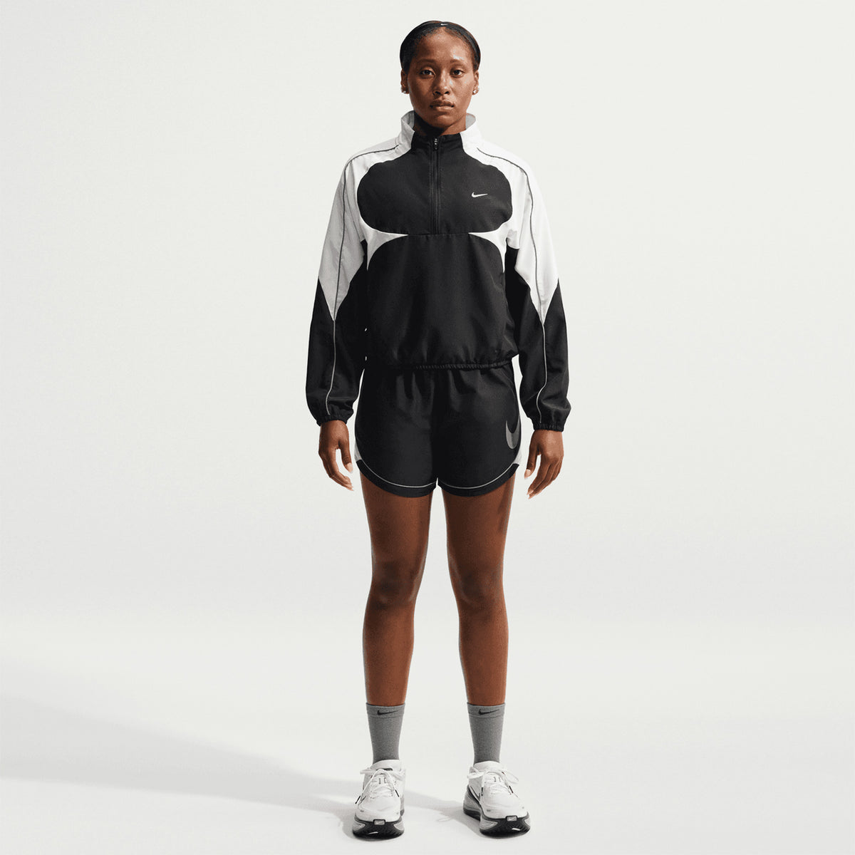 A person stands facing forward wearing a Nike Women’s Dri-FIT Tempo Swoosh 1/2 Zip Running Jacket in Black, White, and Cool Grey against a plain white background.