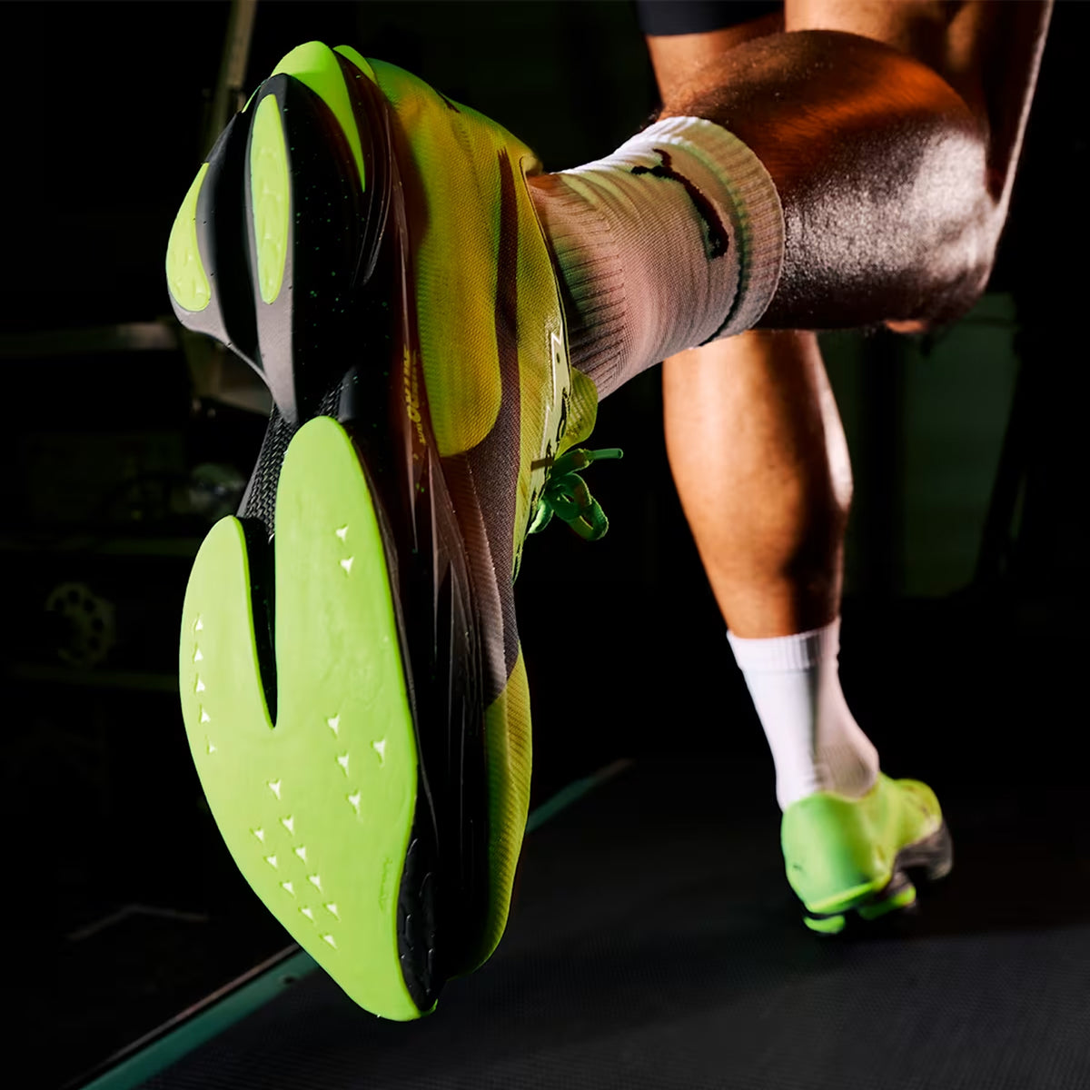 Close-up of a person running on a treadmill, showing the bright green sole of a running shoe and their muscular legs. The individual is wearing white socks.