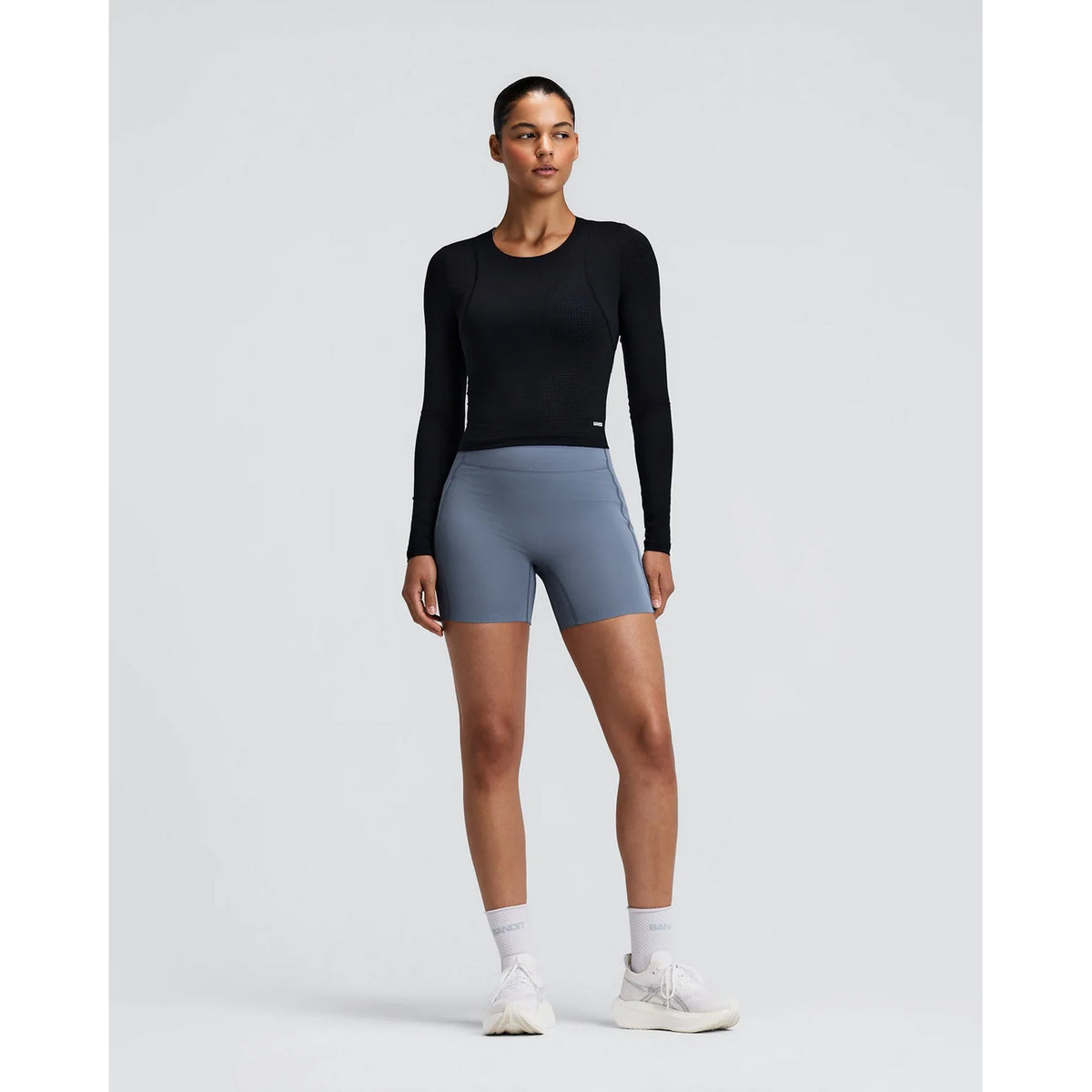 A woman stands against a plain background, wearing the Bandit Women’s GeoMesh Eclipse Long Sleeve Top in black, paired with gray bike shorts, white socks, and white sneakers. She looks forward with a neutral expression and her arms relaxed at her sides.