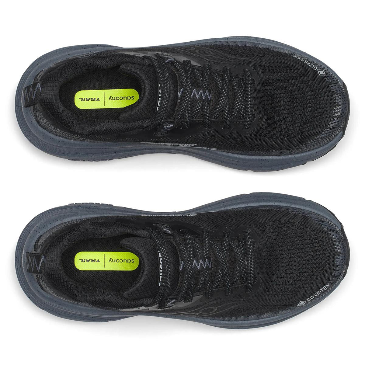 Two black Women’s Guide TR GTX Black / Gunmetal trail running shoes by Saucony with dark soles, shown from above. The yellow insoles read “SUGCONY TRAIL.” The shoes are aligned parallel to each other.