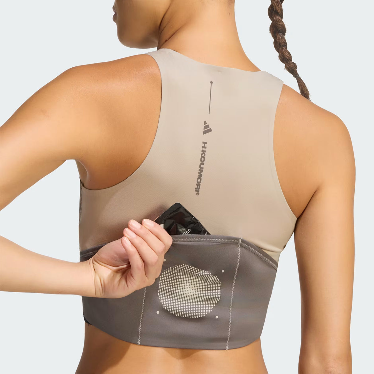 Wearing the Adidas Women’s Hermanos Koumori 1/2 Zip Running Crop in Wonder Beige / Charcoal, a person with a braided hairstyle is shown from the back and arm as they place a black item into the mesh pocket on the gray waistband.