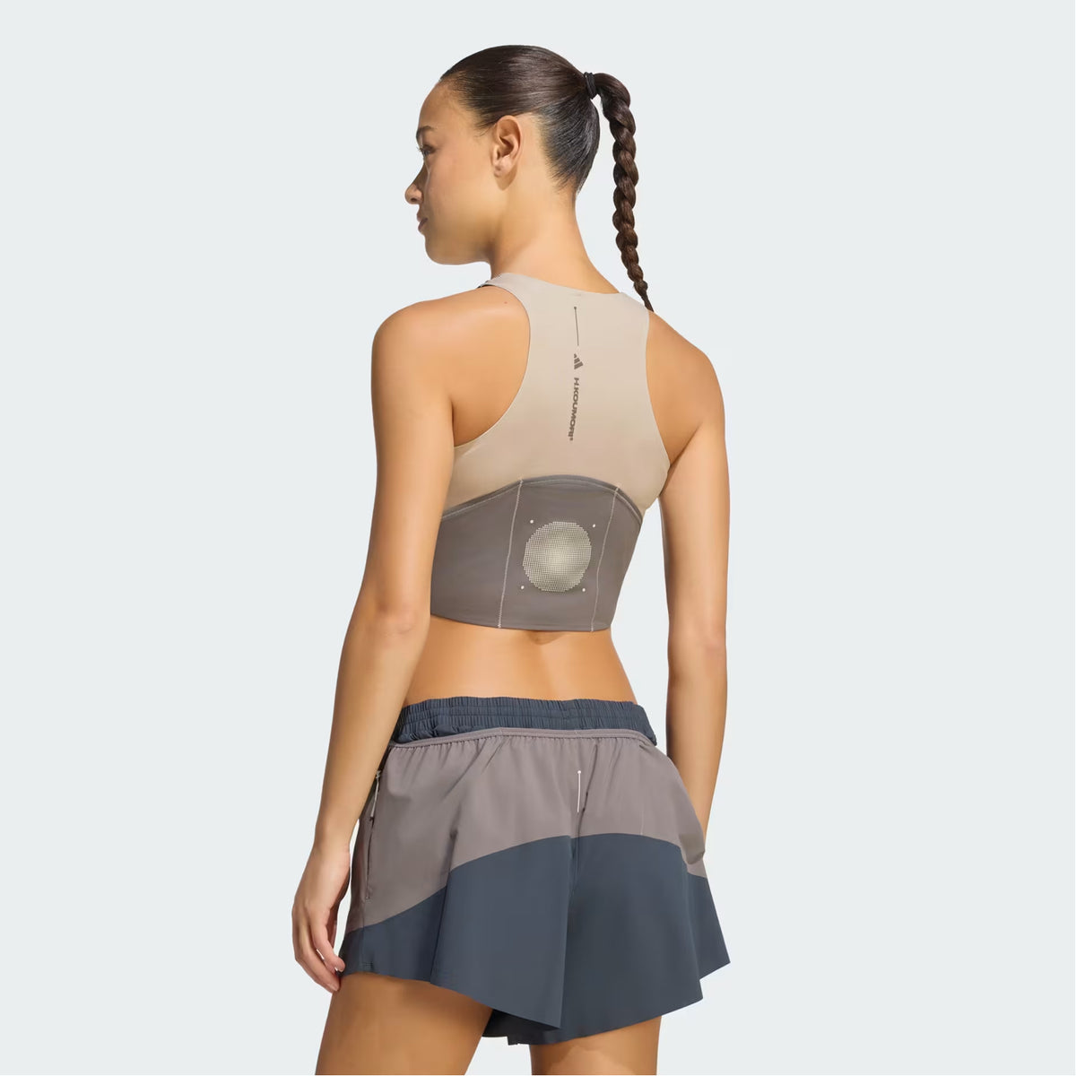 A woman with a long braided ponytail wears the Adidas Women’s Hermanos Koumori 1/2 Zip Running Crop in Wonder Beige and Charcoal, which features a performance fit and a circular device on her back. She pairs it with gray shorts and stands against a light background.
