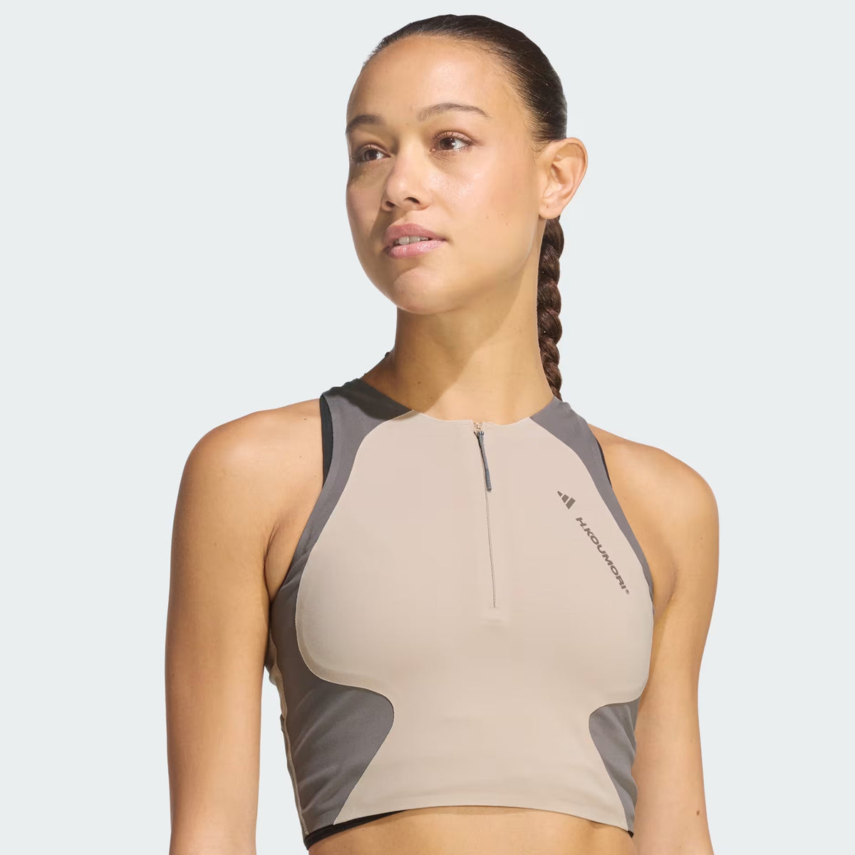 A woman with a long braid wears the Adidas Women’s Hermanos Koumori 1/2 Zip Running Crop in Wonder Beige and Charcoal, featuring a performance fit and sleeveless design, as she looks slightly to the side against a light background.