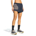 A person wearing Adidas Women’s Hermanos Koumori 3 Inch Running Shorts in Night Grey / Charcoal, featuring a small logo on the gray side pocket, black socks, and gray sneakers stands against a white background. Designed as performance running gear by Adidas, only the lower body is visible.