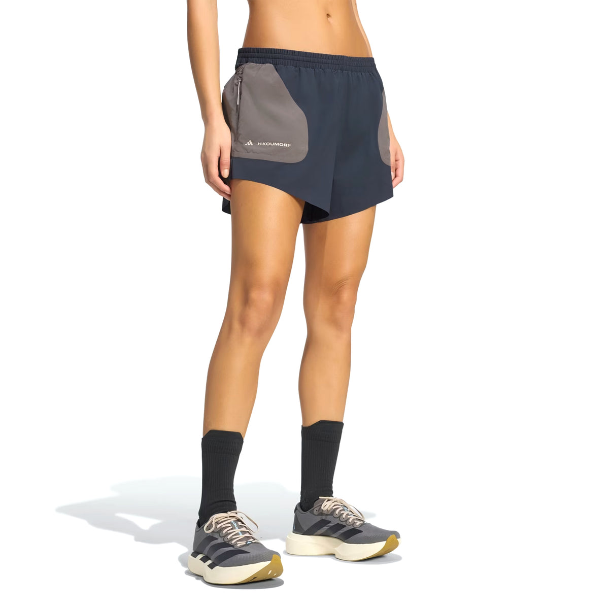 A person wearing Adidas Women’s Hermanos Koumori 3 Inch Running Shorts in Night Grey / Charcoal, featuring a small logo on the gray side pocket, black socks, and gray sneakers stands against a white background. Designed as performance running gear by Adidas, only the lower body is visible.