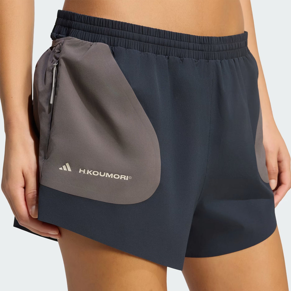 Close-up of a person wearing the Adidas Women’s Hermanos Koumori 3 Inch Running Shorts in Night Grey / Charcoal, featuring a gray zippered pocket on the left side, highlighted with the H.KOUMORI logo and classic Adidas emblem—ideal performance shorts that effortlessly combine running functionality with streetwear appeal.