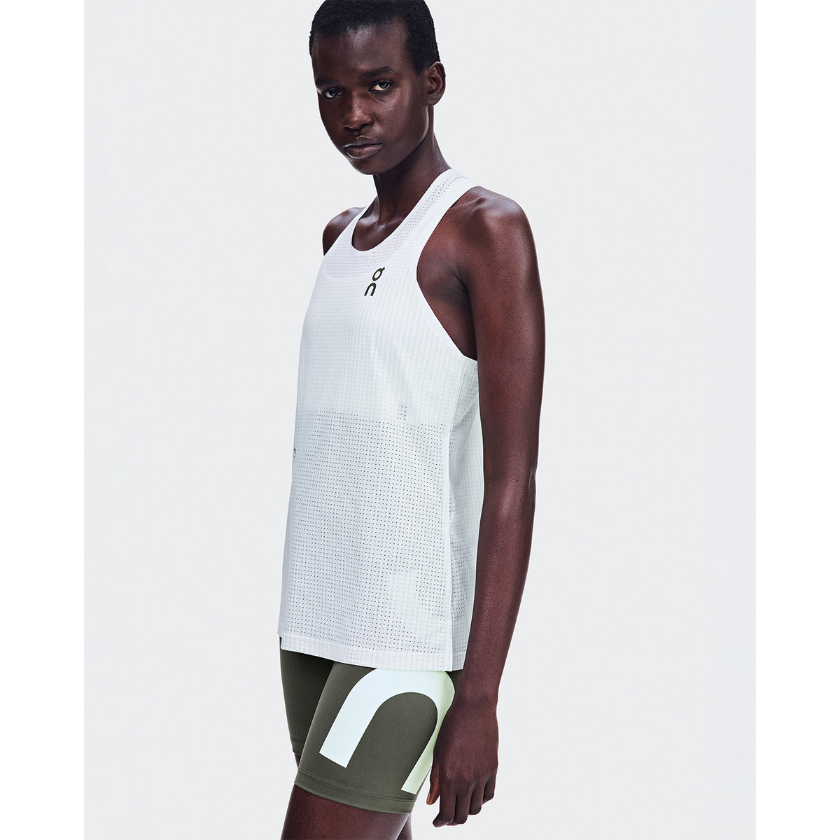 A person wearing a white sleeveless athletic top and olive green shorts with a large white letter design stands against a plain white background, looking toward the camera. The outfit is complemented by the On Women’s Race Half Tights LN1 Mire—form-fitting tights crafted from moisture-wicking fabric—adding both comfort and performance to their athletic style.