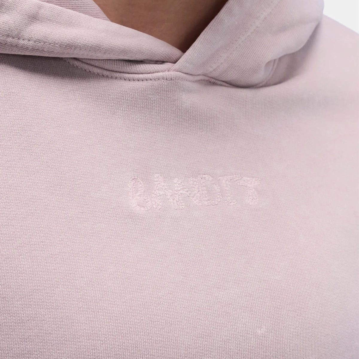 Close-up of a pale pink hoodie with subtle, barely visible embroidered text on the chest, partially obscured by the angle and fabric texture.
