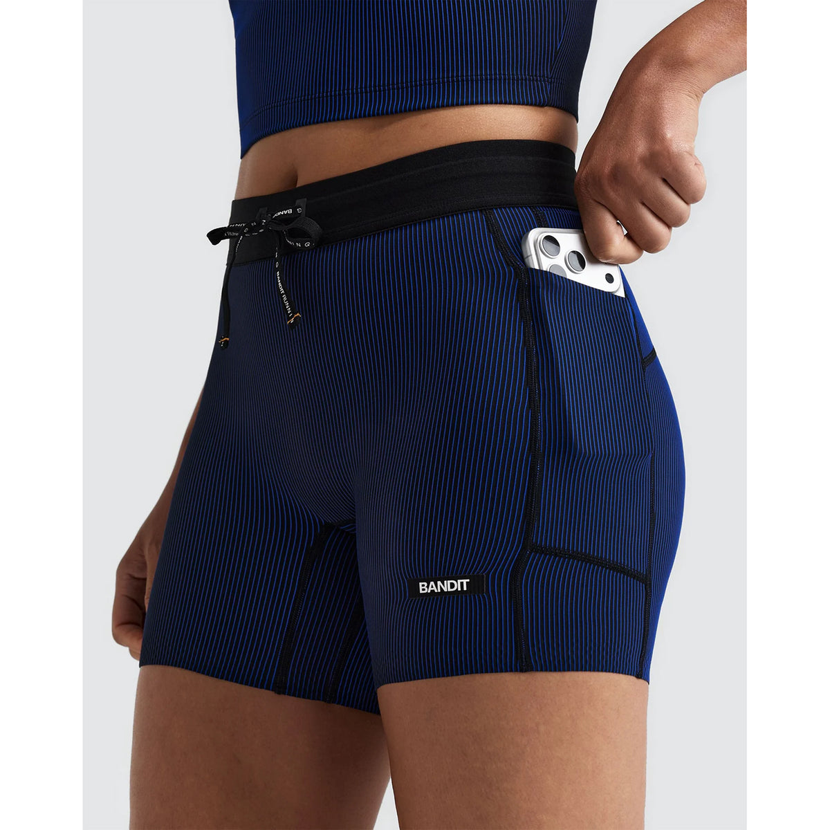 Women’s Striped Cadence 5″ Compression Shorts Black / Bauhaus Blue featuring a black waistband, drawstrings, and a side pocket holding a smartphone.