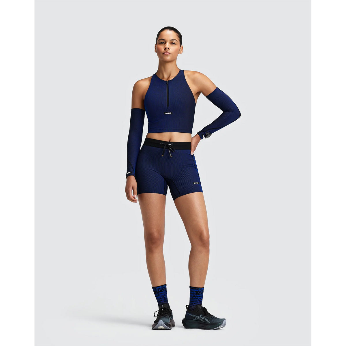 A model wears Women’s Striped Cadence 5″ Compression Shorts Black / Bauhaus Blue with 5-pocket storage, a matching cropped zip-front top with arm sleeves, patterned socks, and running shoes.