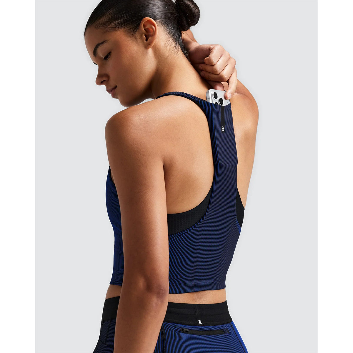 A person wearing a Bandit Women’s Striped Cadence Zip Front T-Back Race Crop in Black / Bauhaus Blue, featuring a blue and black ribbed compression racerback design with a smartphone tucked into a pocket on the back strap.