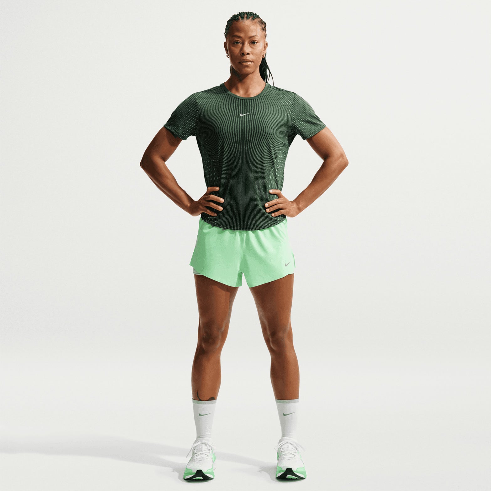 A woman stands with hands on hips, wearing the Nike Women’s Swift Aero-FIT Running Tee in Black / Light Green Spark, paired with light green shorts, white socks, and green-white sneakers against a plain white background.