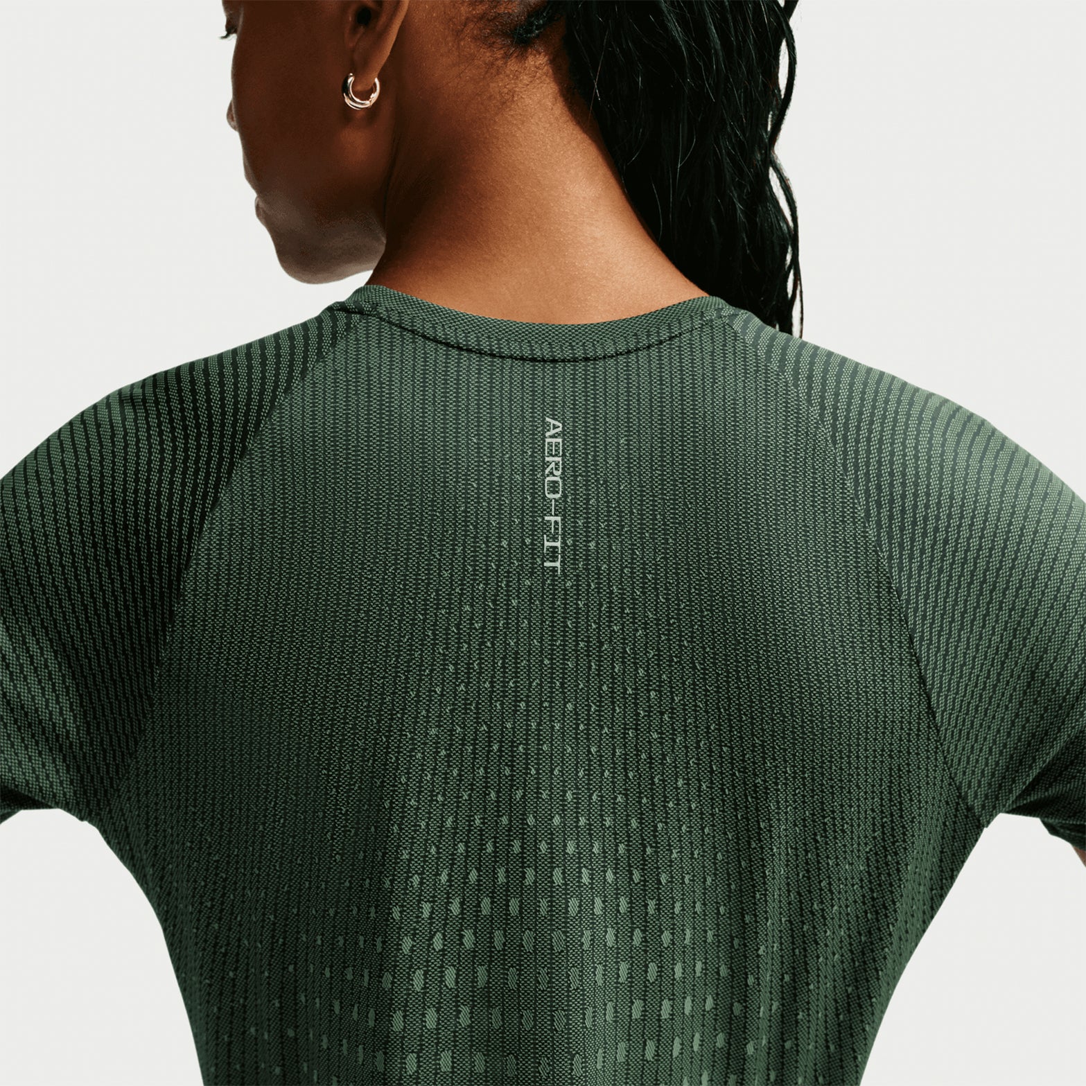 A woman with long dark hair is shown from behind against a plain background, wearing the Nike Women’s Swift Aero-FIT Running Tee in Black / Light Green Spark. The textured, breathable shirt displays the text “AERO FIT” along the upper spine.