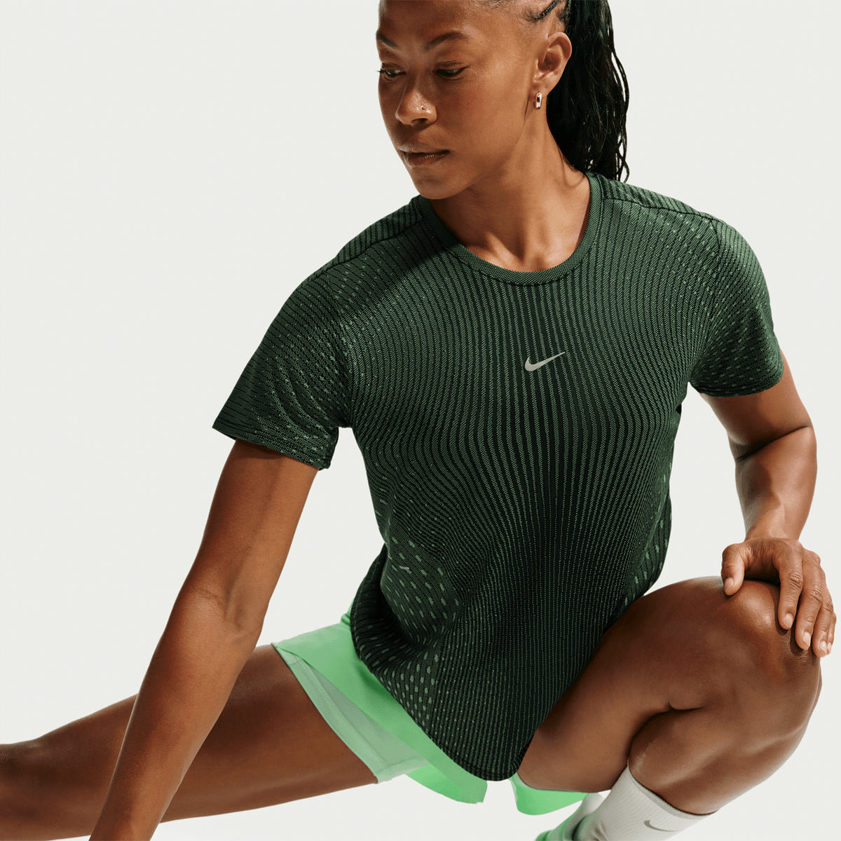 A woman in athletic wear stretches, wearing a dark green Nike shirt and light green shorts, against a plain background.