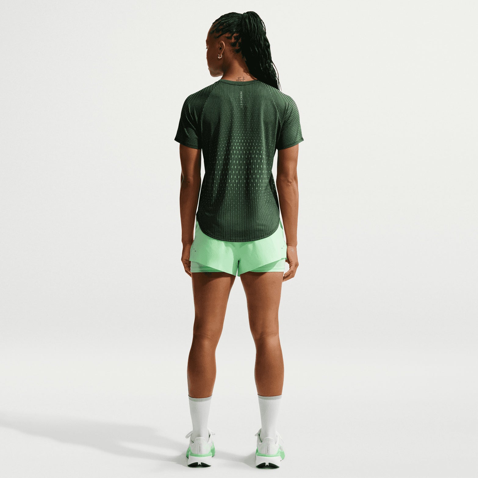 A person with braided hair stands facing away against a plain light background, wearing the Nike Women’s Swift Aero-FIT Running Tee in Black, paired with light green shorts, white socks, and white sneakers.