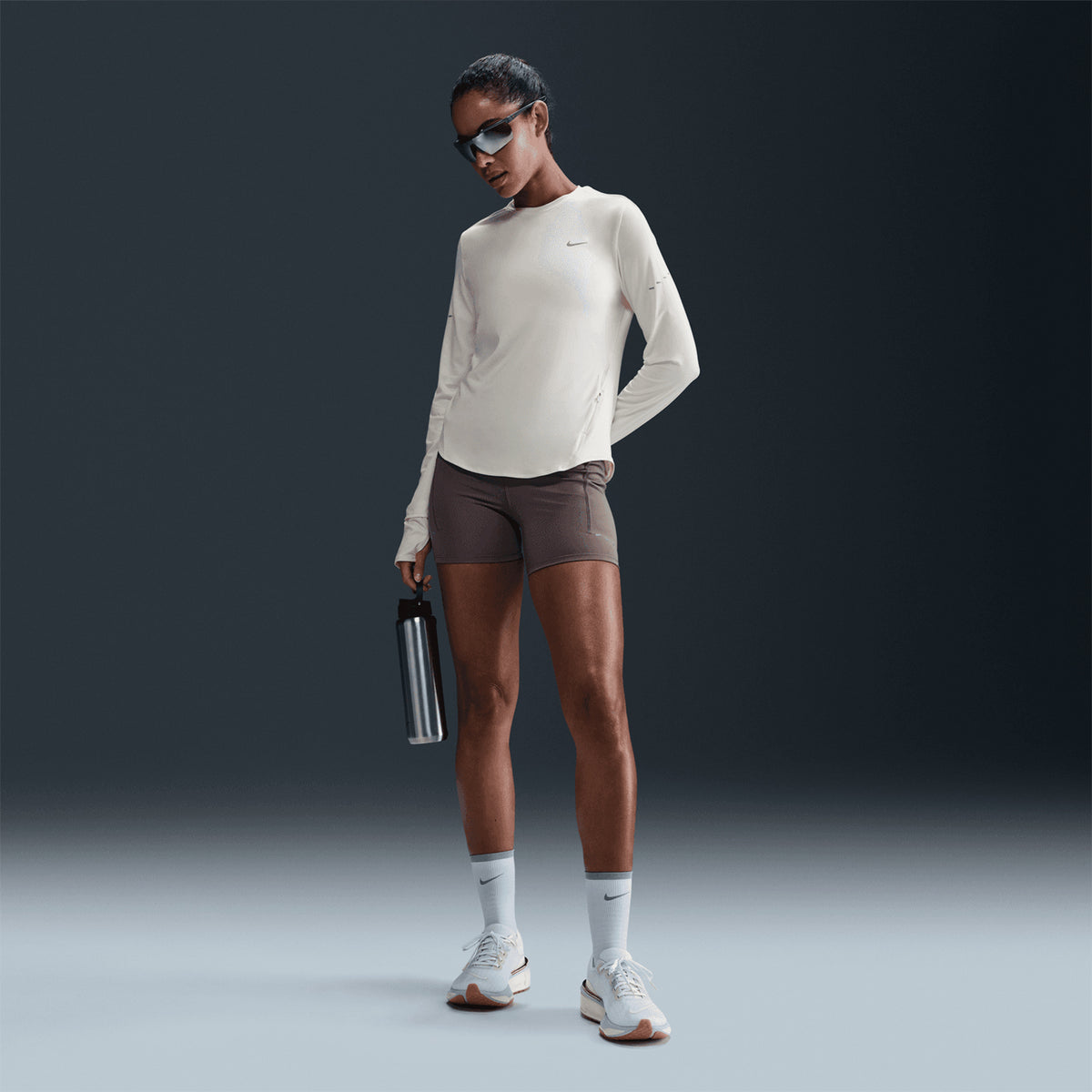 A woman wearing sunglasses and the Nike Women’s Swift Dri-FIT UV Crew-Neck Running Top in Sail, featuring sweat-wicking technology, paired with brown shorts, white socks, and sneakers, holds a water bottle while standing confidently against a dark background.