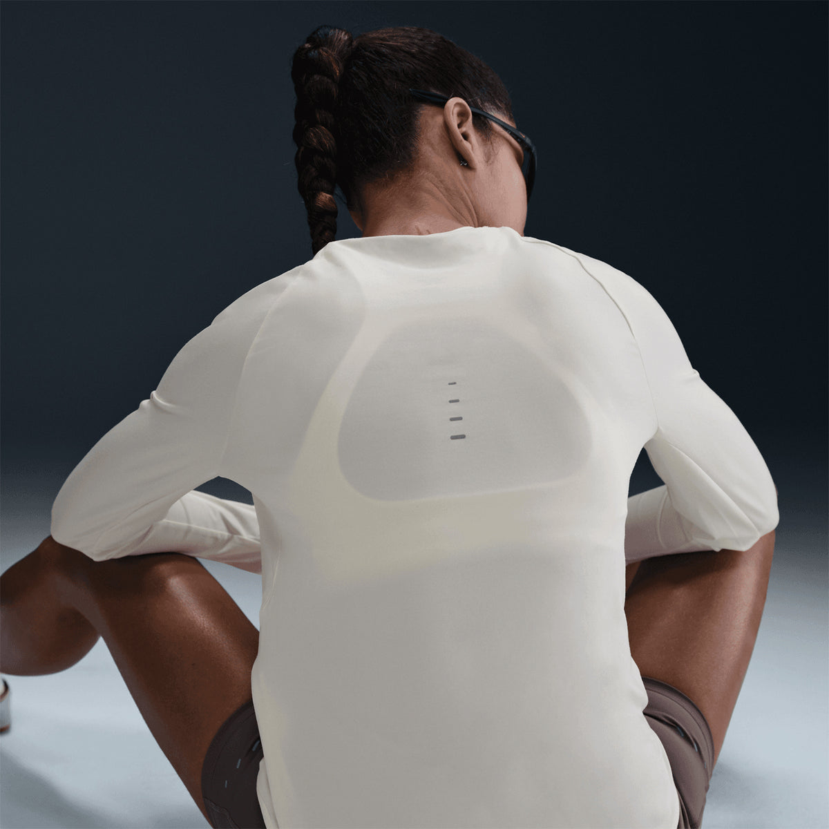A person with a long braid sits on the floor facing away, wearing the Nike Women’s Swift Dri-FIT UV Crew-Neck Running Top in Sail—featuring sweat-wicking fabric and small horizontal lines visible on the back—paired with brown shorts.