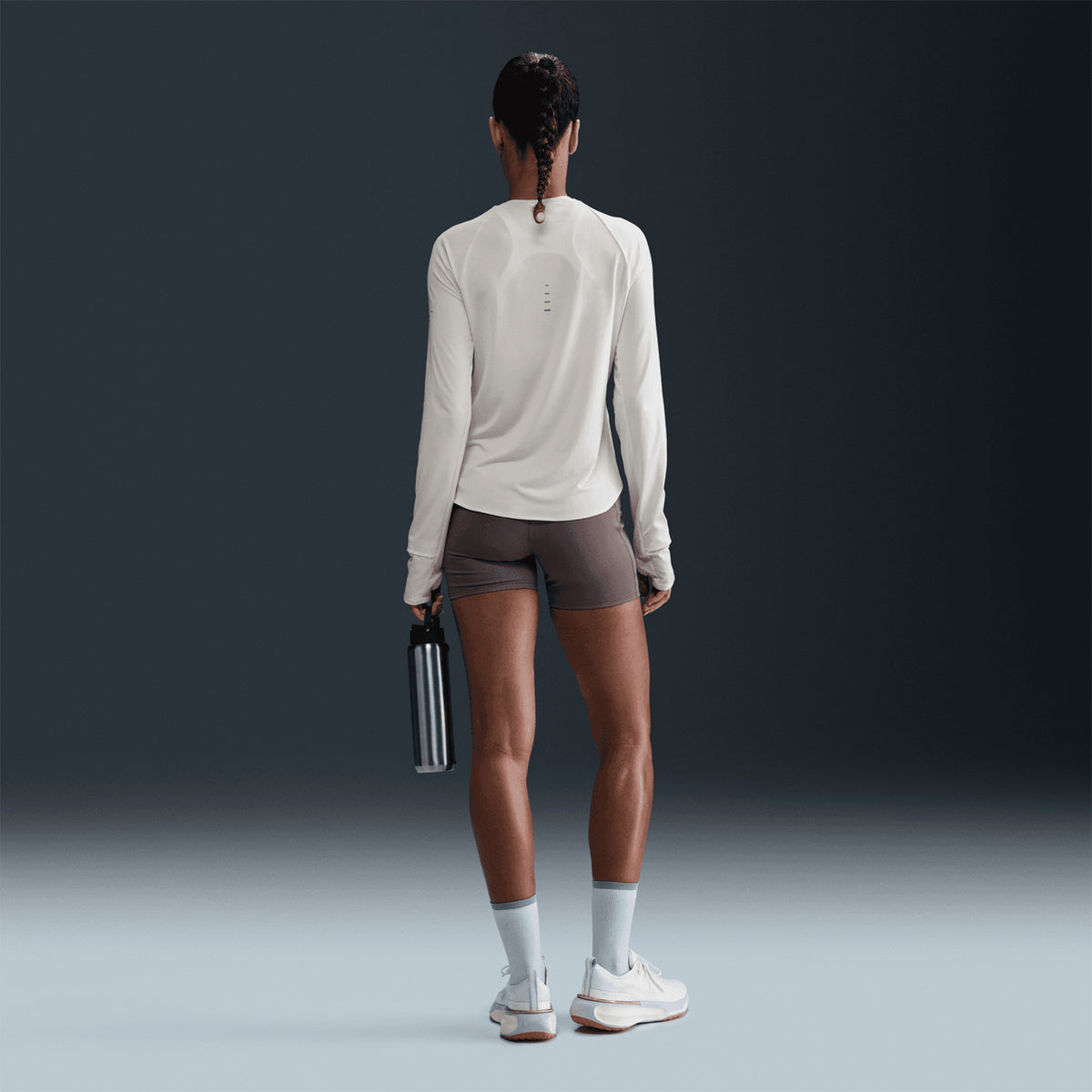 A person with braided hair stands facing away, wearing the Nike Women’s Swift Dri-FIT UV Crew-Neck Running Top in Sail, paired with gray shorts, white socks, and sneakers, and holding a water bottle in their right hand against a dark background.