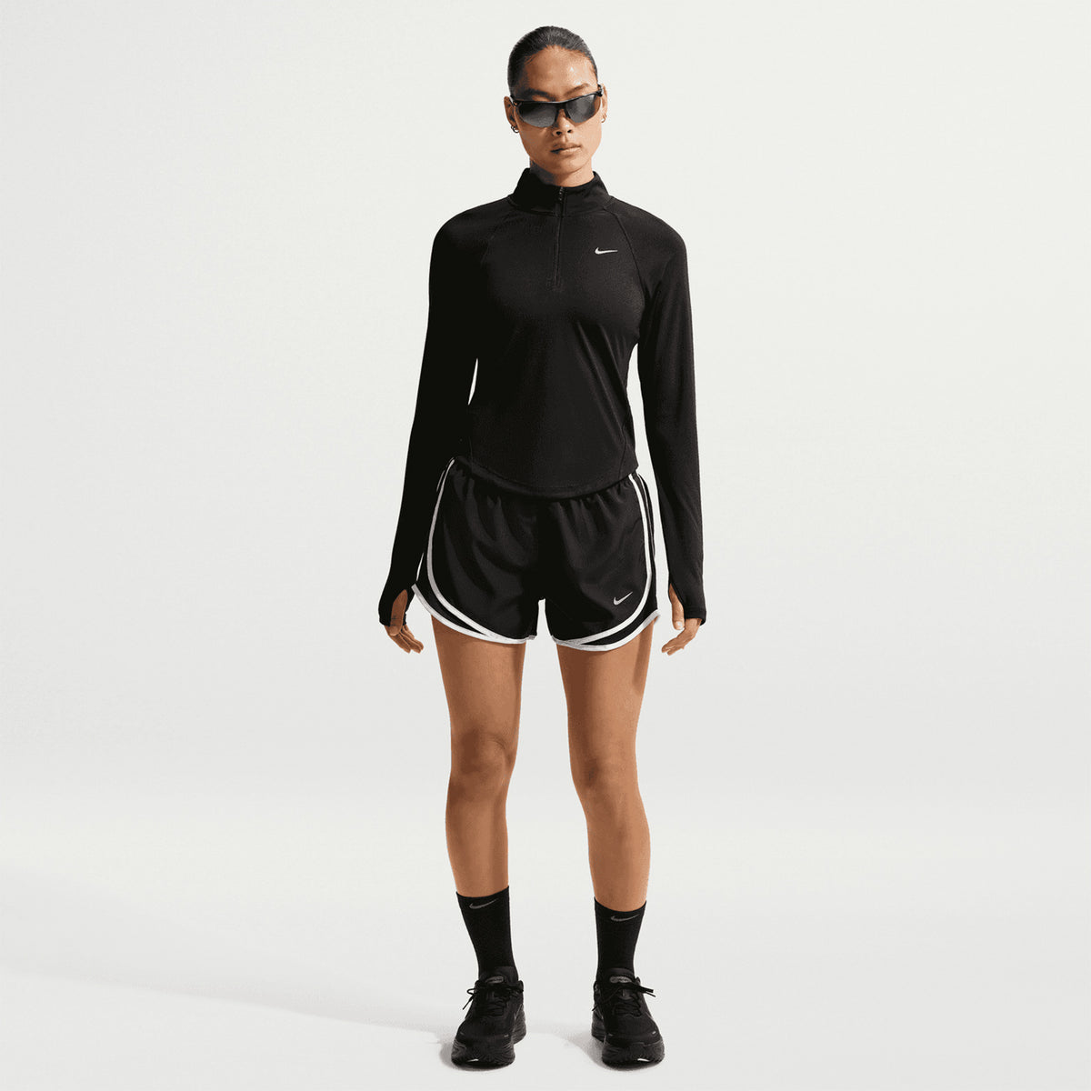 A person stands facing forward against a plain white background, wearing black sunglasses, a black long-sleeve top, black socks, black sneakers, and Nike Women’s Tempo Dri-FIT Mid Rise Running Shorts in Black/Black/White featuring white trim and mesh ventilation for enhanced breathability.
