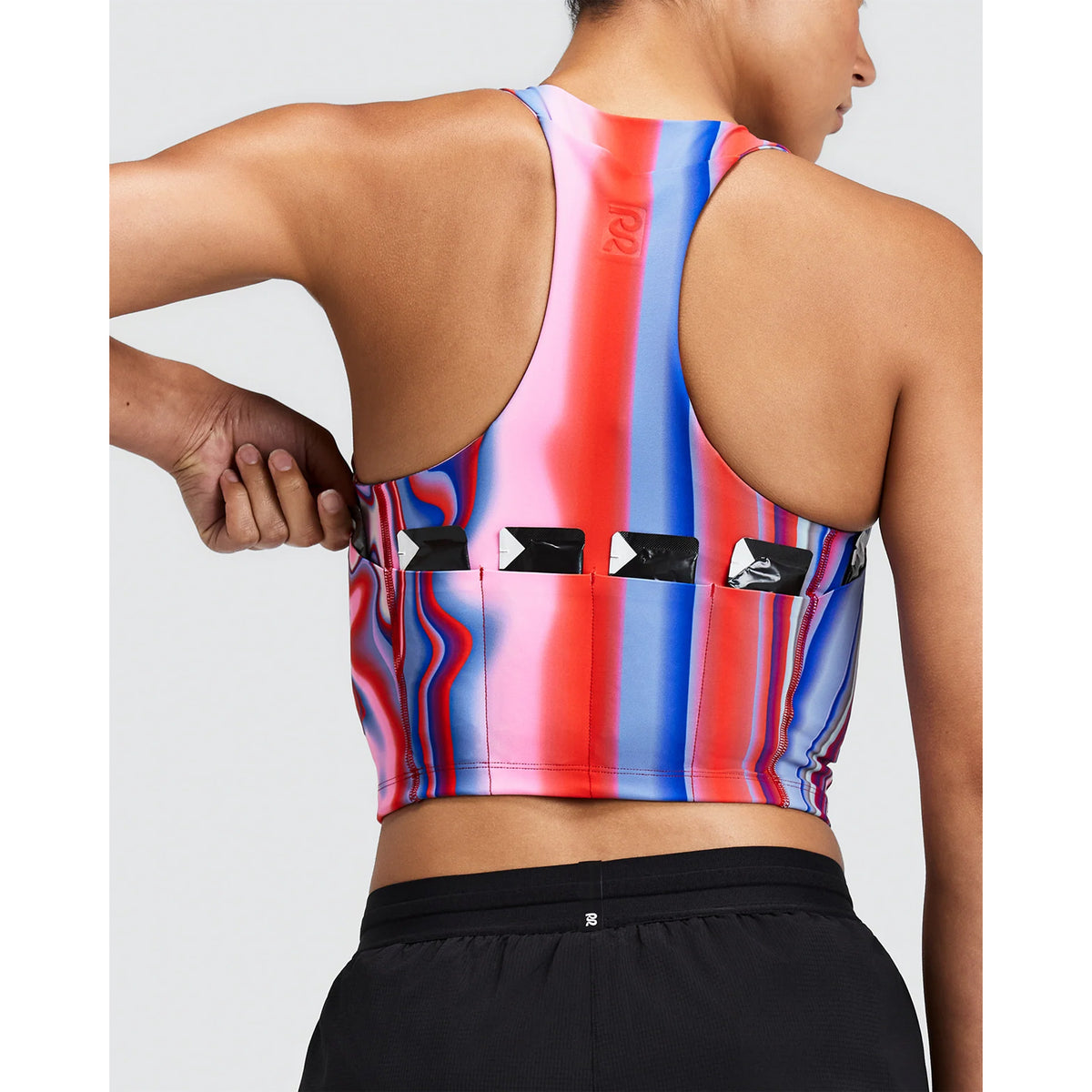 A runner wears a Bandit Women’s Vanisher Race Crop Melted Stripe Gradient compression crop top with a red and blue vertical gradient stripe pattern and a seven-pocket system along the back hem.
