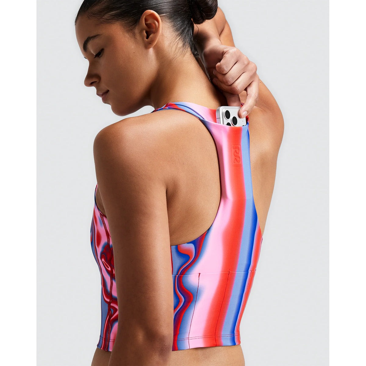 A woman wearing a Bandit Women’s Vanisher Race Crop Melted Stripe Gradient sweat-hiding compression racerback crop top with a pink, blue, and red wavy stripe pattern, putting a smartphone into the pocket on the back strap.